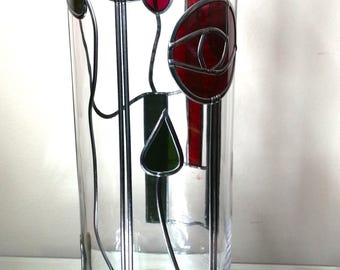 May include: A tall, cylindrical clear glass vase featuring a decorative stained glass overlay. The design includes two prominent red circular floral motifs and a single green leaf shape, all outlined in silver metal solder lines.