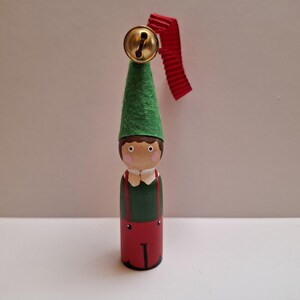 May include: A wooden peg doll ornament featuring a green felt cone hat topped with a gold metal bell and a red ribbon loop. The figure has a painted face with pink cheeks, a green shirt with red suspenders, and red trousers with black details.