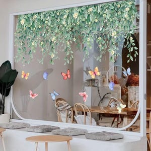 May include: A window decorated with a green and white floral vine design and colorful butterfly decals. The butterflies are in shades of pink, yellow, orange, and blue. The window overlooks a cafe with wooden tables and chairs.