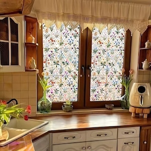 May include: A kitchen window with floral patterned glass, framed by dark wood and a lace valance. The kitchen features white cabinets, a wooden countertop, and a sink with a vase of flowers. The overall style is rustic and charming.