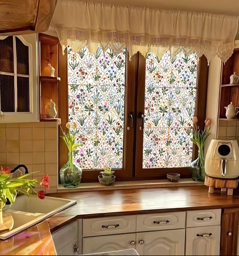 May include: A kitchen window with floral patterned glass, framed by dark wood and a lace valance. The kitchen features white cabinets, a wooden countertop, and a sink with a vase of flowers. The overall style is rustic and charming.