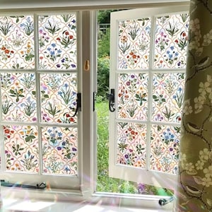 May include: A white window with floral patterned glass panes, displaying a colourful botanical design with red, blue, and purple flowers. A green curtain with bird and floral motifs hangs to the right, with a garden visible outside.