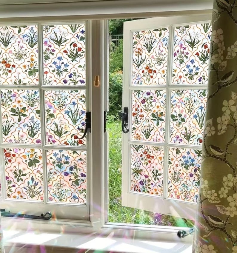 May include: A white window with floral patterned glass panes, displaying a colourful botanical design with red, blue, and purple flowers. A green curtain with bird and floral motifs hangs to the right, with a garden visible outside.