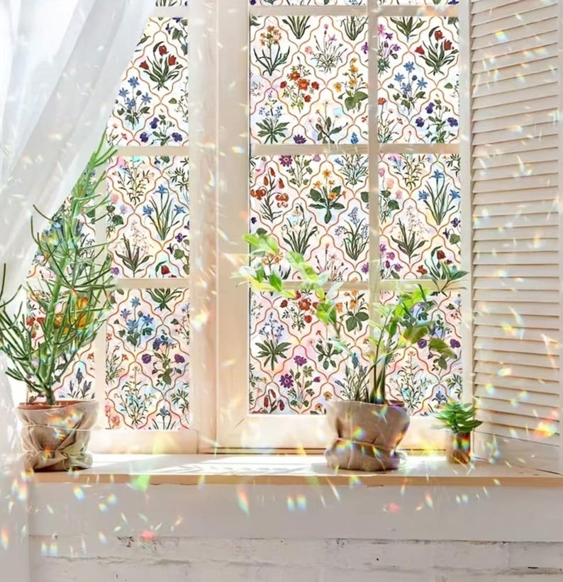 May include: A window with a floral design, showcasing a range of colourful flowers and green leaves on a white backdrop. The window is framed in white, with a wooden sill and potted plants. Sunlight creates a rainbow effect.