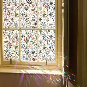 May include: A window featuring a floral design. The window is divided into six sections, each showing a repeating pattern of colourful flowers and green leaves on a white backdrop. The window frame is light-toned, and sunlight passes through, creating a rainbow effect.