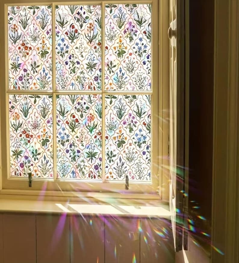 May include: A window featuring a floral design. The window is divided into six sections, each showing a repeating pattern of colourful flowers and green leaves on a white backdrop. The window frame is light-toned, and sunlight passes through, creating a rainbow effect.