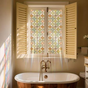 Könnte beinhalten: Ein Badezimmerfenster mit dekorativer Musterfolie. Das Fenster wird von offenen, hellfarbigen Fensterläden flankiert. Die Fensterfolie zeigt ein sich wiederholendes geometrisches Design in Grün-, Gelb-, Orange- und Lilatönen. Eine Badewanne im Vintage-Stil befindet sich im Vordergrund.