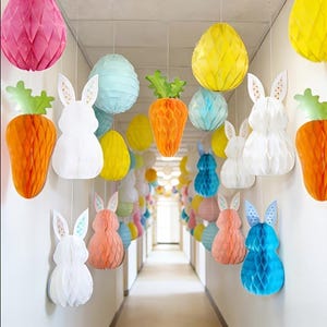May include: A hallway decorated with hanging Easter decorations. The decorations include honeycomb paper carrots, bunny rabbits, and egg-shaped ornaments in various pastel colors. The decorations are suspended from the ceiling.