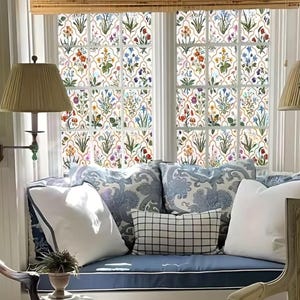 May include: A window seat with floral patterned window film. The film features a repeating pattern of colourful flowers and green leaves. The seat has blue cushions and decorative pillows in white and blue patterns. A bamboo blind is above the window.