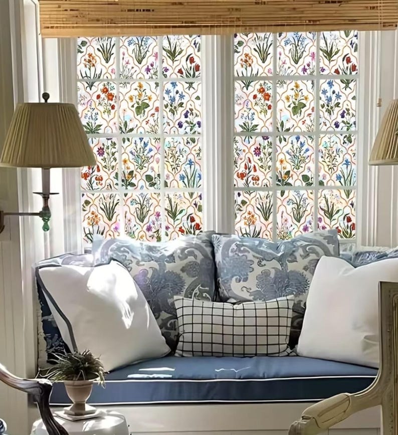 May include: A window seat with floral patterned window film. The film features a repeating pattern of colourful flowers and green leaves. The seat has blue cushions and decorative pillows in white and blue patterns. A bamboo blind is above the window.