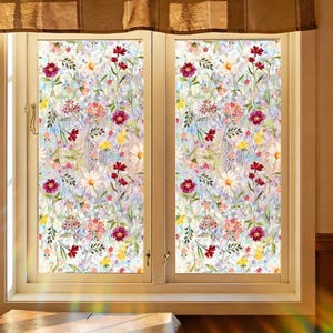 May include: Two-panel window with floral patterned window film. The film features a colourful design of red, pink, yellow, and white flowers with green leaves on a light background. The window frame is white, and a brown curtain is above.