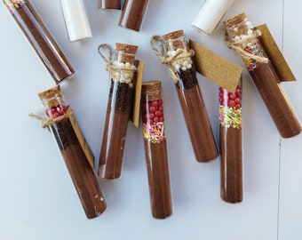 May include: An overhead shot of several glass tubes filled with ingredients for hot chocolate. The tubes have cork stoppers and are tied with twine. Some tubes have tags that read "White Chocolate" and "Hot Chocolate". The tubes contain white powder, chocolate powder, and colorful sprinkles.