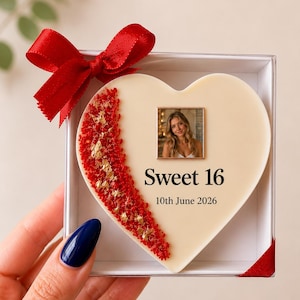 Op de afbeelding: Een hartvormige, crèmekleurige plaat in een witte doos met een rode strik. De plaat bevat een vierkante foto van een persoon, de tekst "Sweet 16" en "10th June 2026", en een decoratieve, gebogen strook van rood en goudkleurig materiaal.