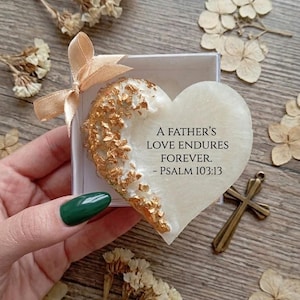 Puede incluir: Un objeto decorativo en forma de corazón con detalles dorados y el texto "A FATHER'S LOVE ENDURES FOREVER. - PSALM 103:13" se presenta en una caja blanca con una cinta color melocotón. También se ve una pequeña cruz de bronce y flores secas.