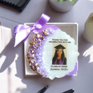 May include: A white, purple, and gold resin geode with a photo of a graduate wearing a cap and gown. The geode has the text "Thank you for celebrating with me. Class of 2025 Jasmine Miller."