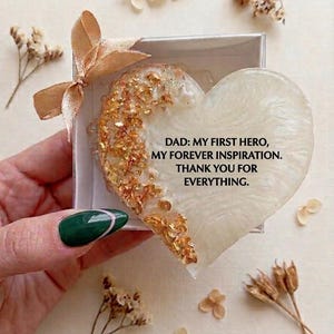 Peut inclure: Un cadeau en forme de cœur dans une boîte avec un ruban doré. Le cœur est blanc avec des paillettes dorées et le texte dit : "DAD: MY FIRST HERO, MY FOREVER INSPIRATION. THANK YOU FOR EVERYTHING."