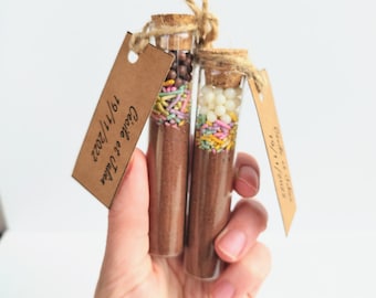 May include: Two small glass vials filled with brown powder and colorful sprinkles, sealed with corks and tied with twine. Each vial has a small brown tag with handwritten text. The vials are held in a hand.