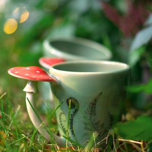 Può includere: Due tazze in ceramica verde con manici a forma di fungo e cime rosse con pois bianchi. Le tazze sono decorate con illustrazioni di felci e si trovano nell'erba verde, creando un tema stravagante ispirato alla natura.
