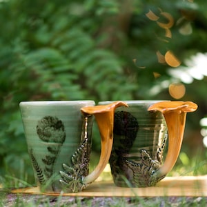 Può includere: Due tazze in ceramica con smalto verde e manici a forma di fungo. Le tazze presentano disegni di felci e funghi. I manici sono di colore arancione caldo. Le tazze sono su una superficie di legno.
