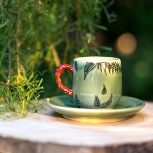 Op de afbeelding: Een kleine, lichtgroene keramische koffiekop en schotel set. De beker heeft een rode handgreep met witte stippen en is versierd met zwarte varenbladmotieven. De schotel heeft dezelfde kleur als de beker. De set staat op een houten oppervlak.