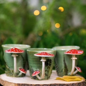 Può includere: Quattro tazze in ceramica verde con dettagli di funghi rossi e bianchi. Le tazze sono decorate con disegni di felci e sono appoggiate su una fetta di legno. Lo sfondo è verde sfocato con bokeh.