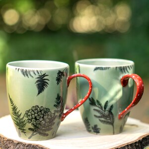 Op de afbeelding: Twee groene keramische mokken met rode stippen handvatten. Elke mok heeft zwarte botanische illustraties van varens en bessen. De mokken worden gepresenteerd op een houten schijf, met een vage groene achtergrond.