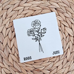 Könnte beinhalten: Eine weiße quadratische Karte mit einer schwarzen Strichzeichnung von drei Rosen. Auf der Karte steht links unten das Wort "ROOS" und rechts unten "Juni". Die Karte liegt auf einem gewebten, hellbraunen Hintergrund.