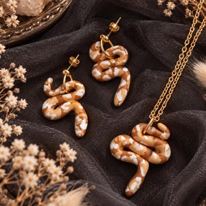 May include: A jewelry set consisting of two stud earrings and one pendant necklace, each featuring a coiled snake charm with a mottled pattern of brown, tan, and white colors, attached to gold-toned metal hardware and a delicate chain.