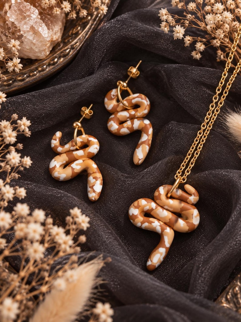 May include: A jewellery set consisting of two stud earrings and one pendant necklace, each featuring a coiled snake charm with a mottled pattern of brown, tan, and white colours, attached to gold-toned metal hardware and a delicate chain.