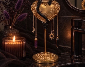 May include: A gold-coloured metal stand featuring a central heart-shaped ornament topped with a flame motif and wrapped in a black snake. Several thin gold chains hang from the heart, displaying charms including a crescent moon, a sun, and two crystal pendants.