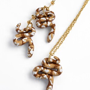 May include: A jewellery set consisting of two snake-shaped earrings and a matching snake pendant on a gold-tone chain. Each snake figure features a brown, tan, and white mottled pattern with a smooth finish and gold-tone metal hardware components.