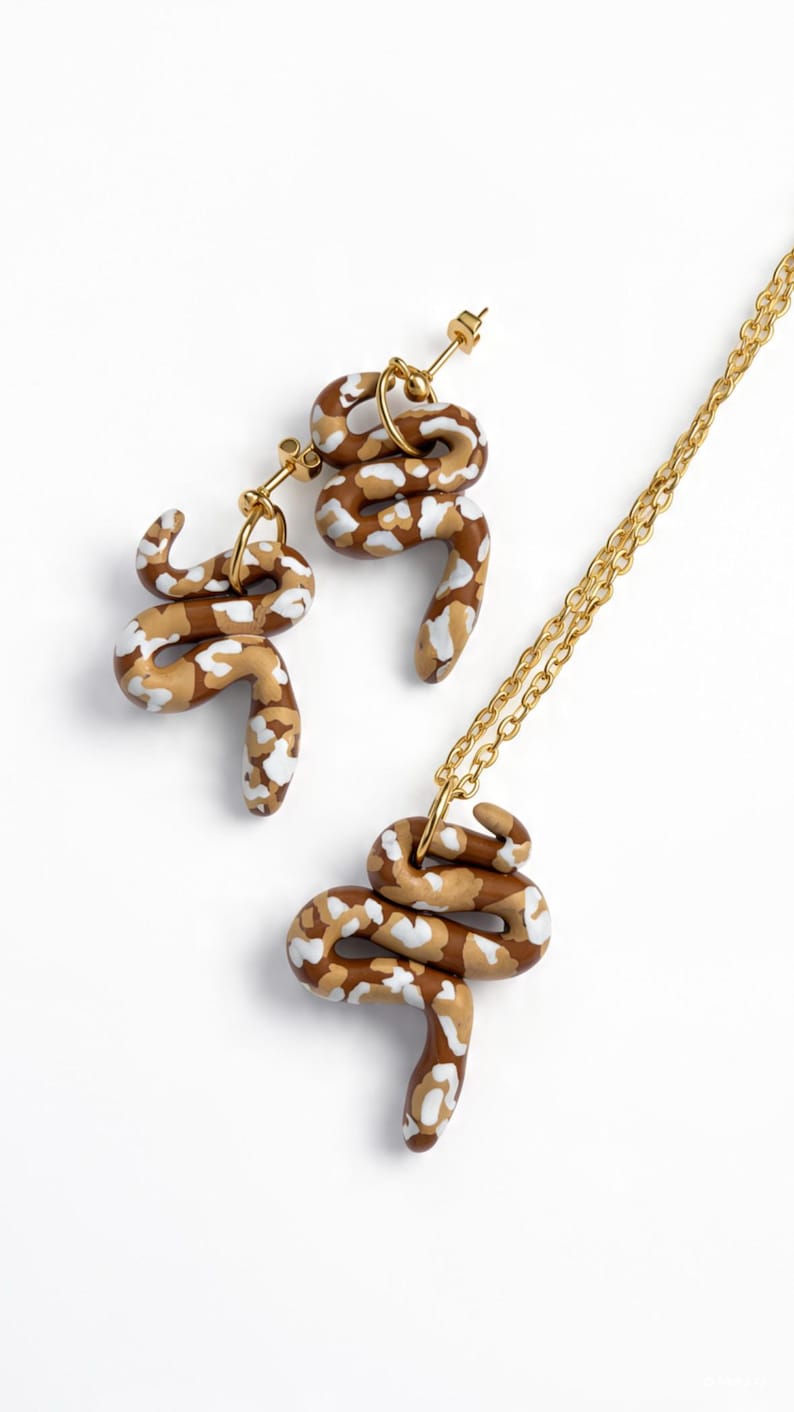 May include: A jewellery set consisting of two snake-shaped earrings and a matching snake pendant on a gold-tone chain. Each snake figure features a brown, tan, and white mottled pattern with a smooth finish and gold-tone metal hardware components.