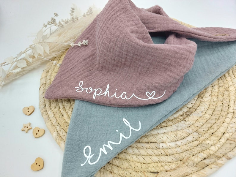 May include: Two personalized muslin baby bibs, one in dusty rose with the name "Sophia" and a heart, and one in light blue with the name "Emil".