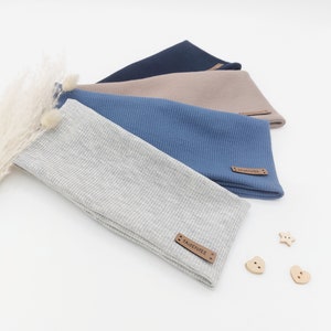 Könnte beinhalten: Vier Stirnbänder aus Rippstrick in Grau-, Blau-, Beige- und Marineblautönen werden gezeigt. Jedes Stirnband hat ein kleines Lederetikett mit dem Text "FADENIEE". Ein Stirnband hat einen kleinen Bommel. Holzknöpfe in Form von Herzen und einem Stern sind in der Nähe verstreut.