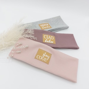 May include: Three fabric headbands with different colors and text on the front. The top headband is gray with the text "FRECH Dachs". The middle headband is pink with the text "CIAO kakao". The bottom headband is light pink with the text "stay COOL".