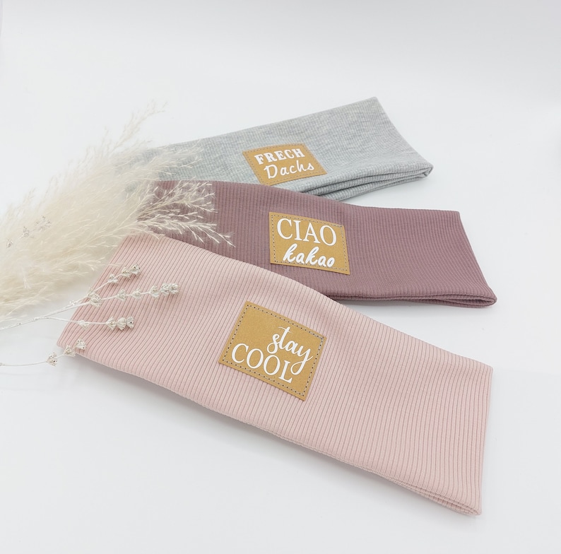 May include: Three fabric headbands with different colors and text on the front. The top headband is gray with the text "FRECH Dachs". The middle headband is pink with the text "CIAO kakao". The bottom headband is light pink with the text "stay COOL".