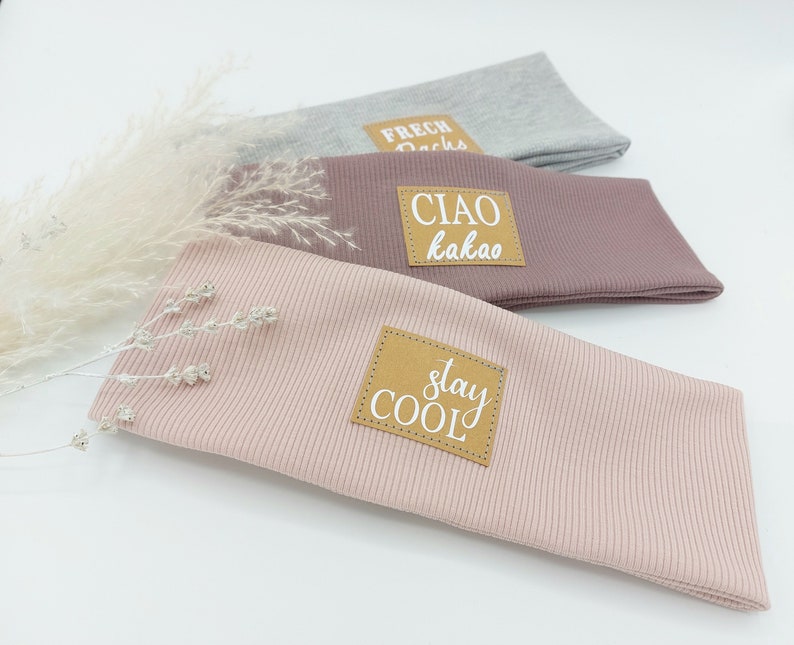 May include: Three fabric headbands with different colors and phrases. The top headband is gray with the phrase "Frech Dachs". The middle headband is pink with the phrase "Ciao kakao". The bottom headband is light pink with the phrase "Stay Cool".