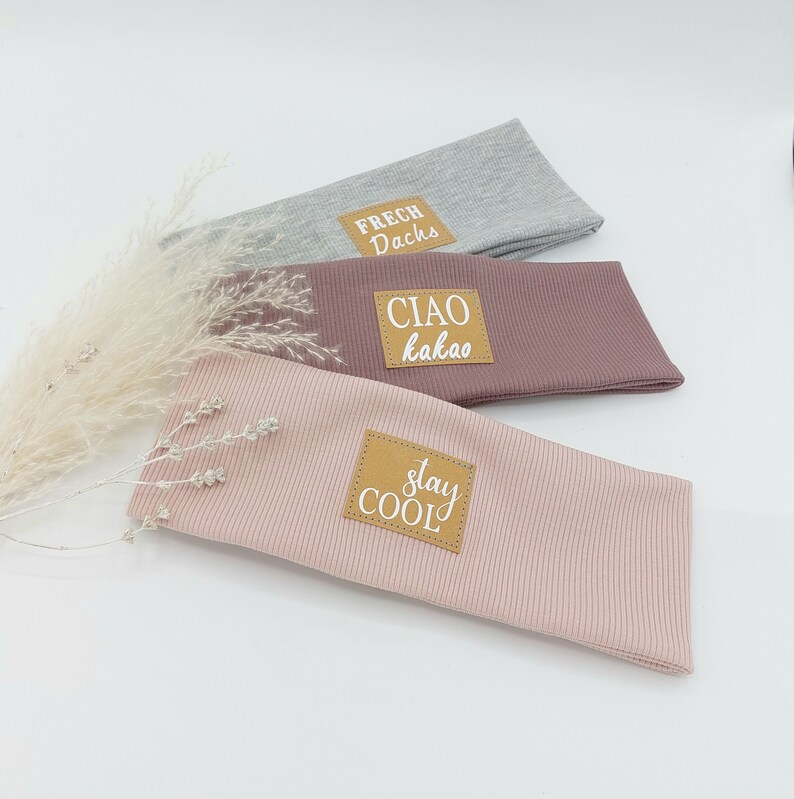 May include: Three fabric headbands with brown leather patches. The top headband is gray with the text "FRECH Dachs". The middle headband is pink with the text "CIAO kakao". The bottom headband is light pink with the text "stay COOL".