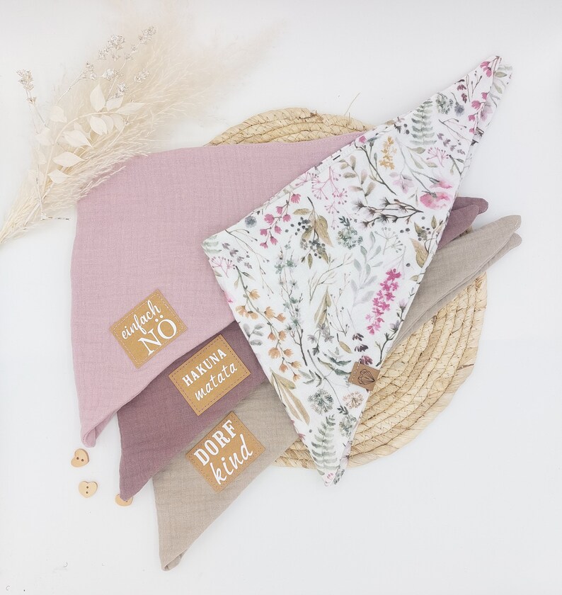 May include: Three fabric bandanas with different patterns and colors. The top bandana is white with a floral pattern, the middle bandana is pink, and the bottom bandana is beige. Each bandana has a brown leather patch with white text. The top bandana says "Hakuna Matata", the middle bandana says "Einfach No", and the bottom bandana says "Dorf Kind".