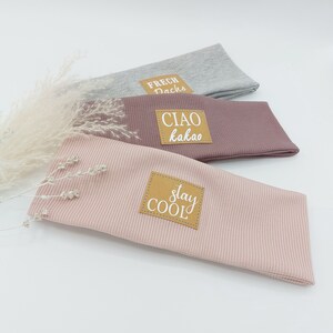 May include: Three fabric headbands with different colors and text on brown patches. The top headband is gray with the text "FRECH DACHs". The middle headband is pink with the text "CIAO kakao". The bottom headband is light pink with the text "stay COOL".