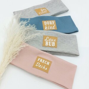 May include: Four gray, blue, and pink headbands with brown leather patches. The patches have white text that reads "Stay Cool", "Dorf Kind", "Laus Bub", and "Frech Dachs".