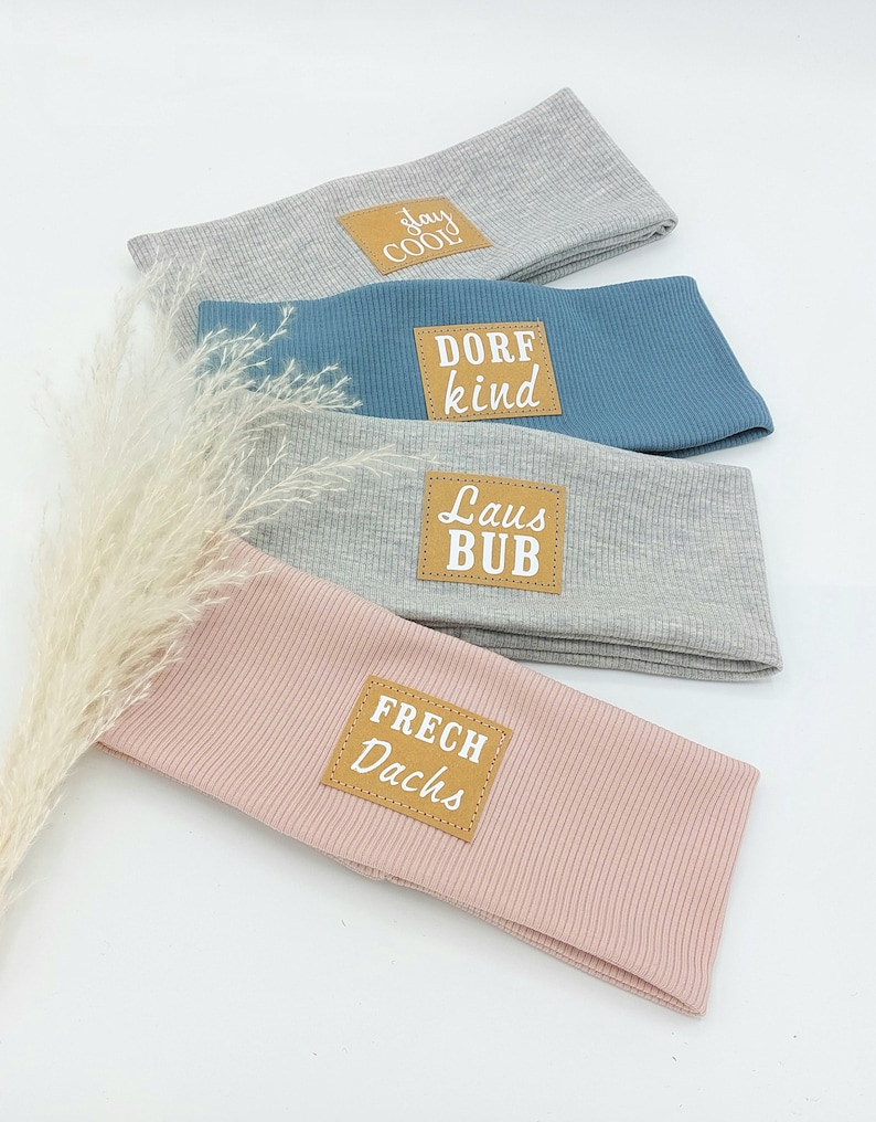 May include: Four gray, blue, and pink headbands with brown leather patches. The patches have white text that reads "Stay Cool", "Dorf Kind", "Laus Bub", and "Frech Dachs".