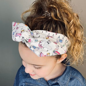 May include: A white headband with a colorful llama print. The headband is tied in a knot on top of the head.