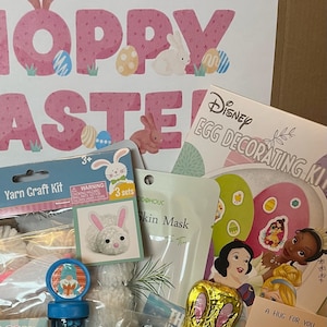 May include: A gift box filled with Easter-themed items, including a yarn craft kit, a bunny-shaped soap, jelly beans, a bubble wand, a chocolate bunny, and plastic Easter eggs in various colors. The box is decorated with a pink and white "Hoppy Easter" sign and a Disney egg decorating kit.