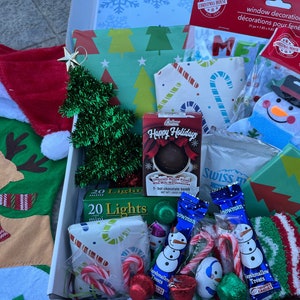 K&ouml;nnte beinhalten: Eine wei&szlig;e Box gef&uuml;llt mit weihnachtlichen Artikeln, darunter ein roter Strumpf mit einem Rentiermotiv, ein kleiner gr&uuml;ner Lametta-Weihnachtsbaum, eine Packung mit 20 Mini-Lichtern, ein Schneemann-Anh&auml;nger, eine Filz-Girlande, eine hei&szlig;e Schokoladenbombe und eine Packung Swiss Miss hei&szlig;e Schokolade.