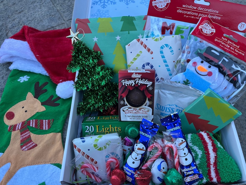 K&ouml;nnte beinhalten: Eine wei&szlig;e Box gef&uuml;llt mit weihnachtlichen Artikeln, darunter ein roter Strumpf mit einem Rentiermotiv, ein kleiner gr&uuml;ner Lametta-Weihnachtsbaum, eine Packung mit 20 Mini-Lichtern, ein Schneemann-Anh&auml;nger, eine Filz-Girlande, eine hei&szlig;e Schokoladenbombe und eine Packung Swiss Miss hei&szlig;e Schokolade.