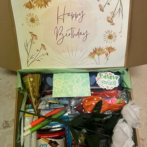 K&ouml;nnte beinhalten: Ein brauner Karton, gef&uuml;llt mit Geburtstagsgeschenken. Die Schachtel ist mit einem Blumendesign und den Worten "Happy Birthday" verziert. Zu den Geschenken geh&ouml;ren Ballons, eine Partym&uuml;tze, Marker, ein Glas Nutella und eine Karte mit der Aufschrift "Glaube an dich selbst".