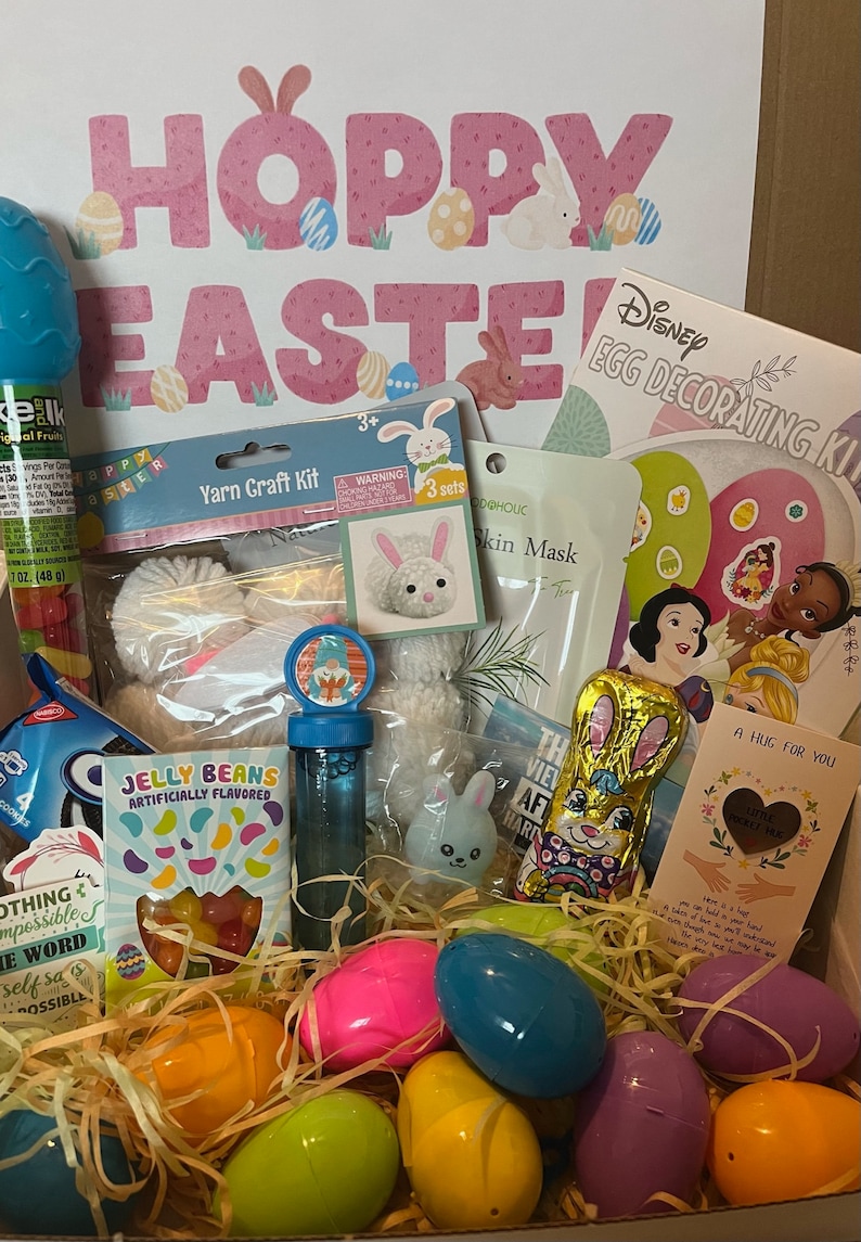 K&ouml;nnte beinhalten: Eine Geschenkbox gef&uuml;llt mit Ostern-Artikeln, darunter ein Garn-Bastelset, eine seifenf&ouml;rmige Hasenfigur, Gummib&auml;rchen, eine Blasenstab, ein Schokoladenhase und Plastik-Ostereier in verschiedenen Farben. Die Box ist mit einem rosa und wei&szlig;en Schild "Hoppy Easter" und einem Disney-Eier-Dekorationsset verziert.