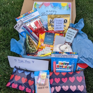 K&ouml;nnte beinhalten: Eine blaue und wei&szlig;e Geschenkbox gef&uuml;llt mit Geburtstags-Partyzubeh&ouml;r, darunter ein Buchstabenbanner, Kerzen, Konfetti und ein Zettel mit der Aufschrift "Happy Birthday".