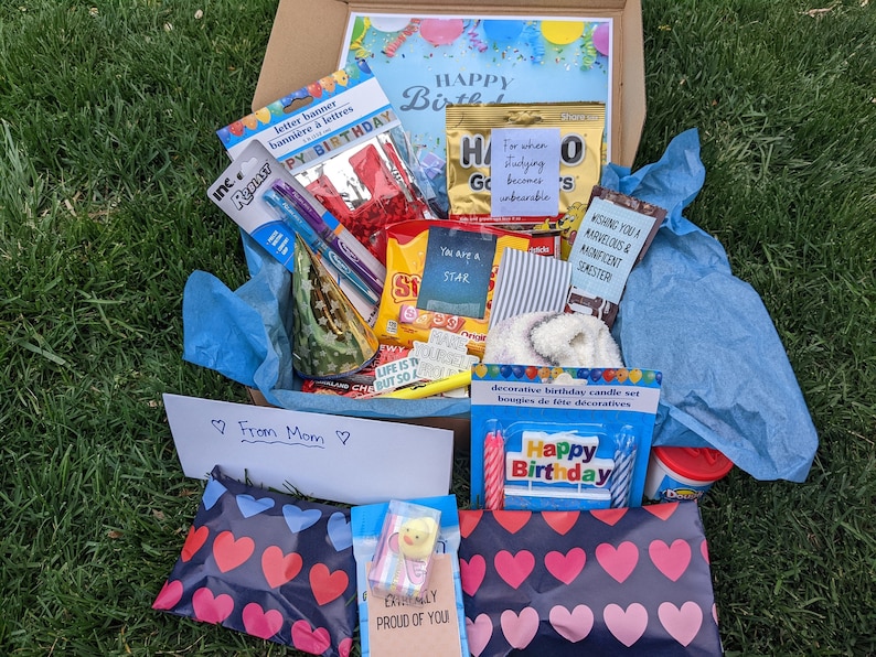K&ouml;nnte beinhalten: Eine blaue und wei&szlig;e Geschenkbox gef&uuml;llt mit Geburtstags-Partyzubeh&ouml;r, darunter ein Buchstabenbanner, Kerzen, Konfetti und ein Zettel mit der Aufschrift "Happy Birthday".